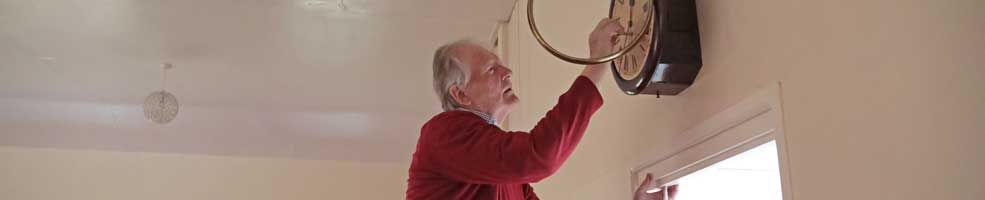 Mervyn Lack winding the clock in Hatley Village Hall – Mervyn had it repaired after years of it not going properly (most not at all!) on 14th July 2022 – from memories of Hatley, by Mervyn Lack, July 2022. Photo: Peter Mann.
